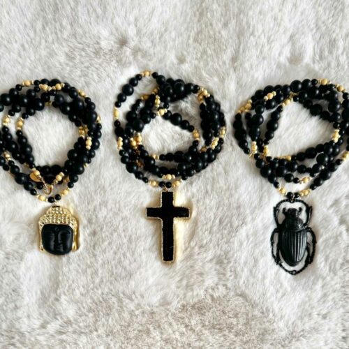Trois sautoirs NOX en perles d’onyx noir mat et détails dorés, avec pendentifs Bouddha, croix noire et scarabée, présentés sur un fond clair.
