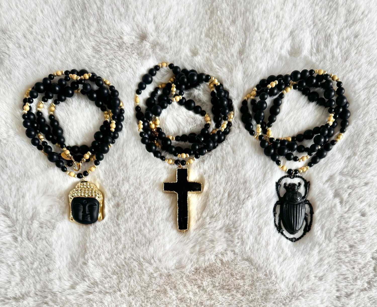 NOX-collection-drop-02-sautoirs-bouddha-croix-scarabee-onyx-noir-mademoiselle-oups Trois sautoirs NOX en perles d’onyx noir mat et détails dorés, avec pendentifs Bouddha, croix noire et scarabée, présentés sur un fond clair.