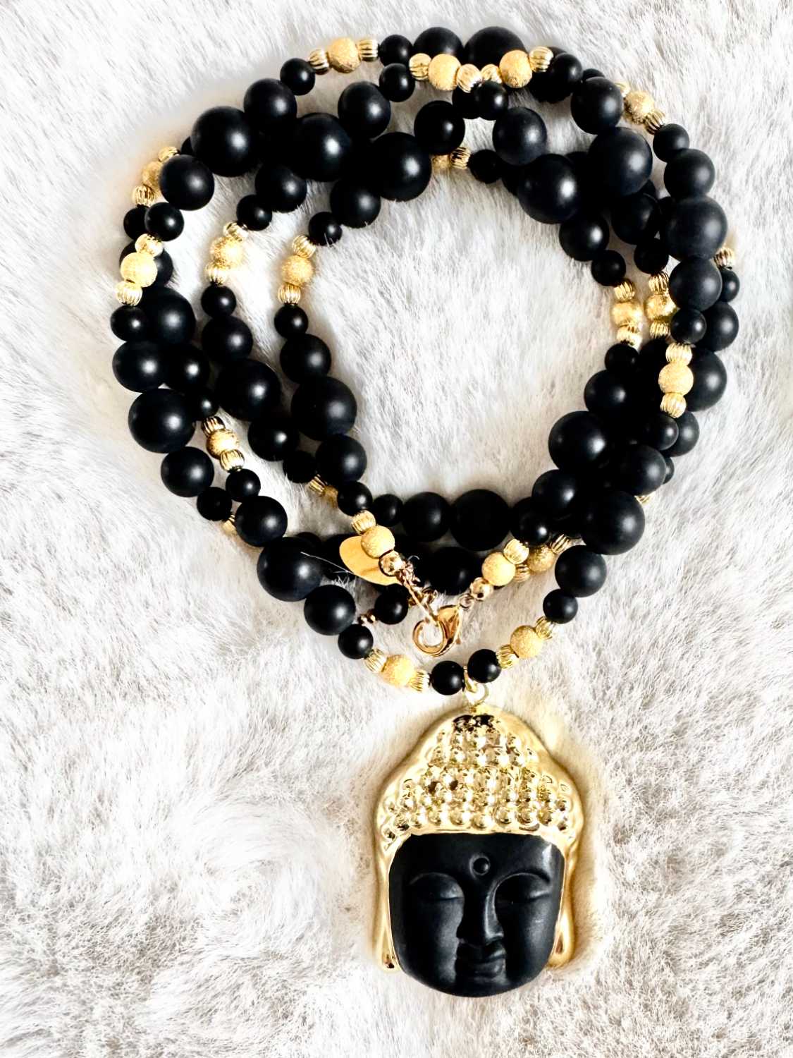 NOX-sautoir-bouddha-noir-onyx-mat-details-dores-mademoiselle-oups Sautoir NOX en perles d’onyx noir mat de différentes tailles, perles dorées et pendentif Bouddha noir et or, présenté sur fond clair.