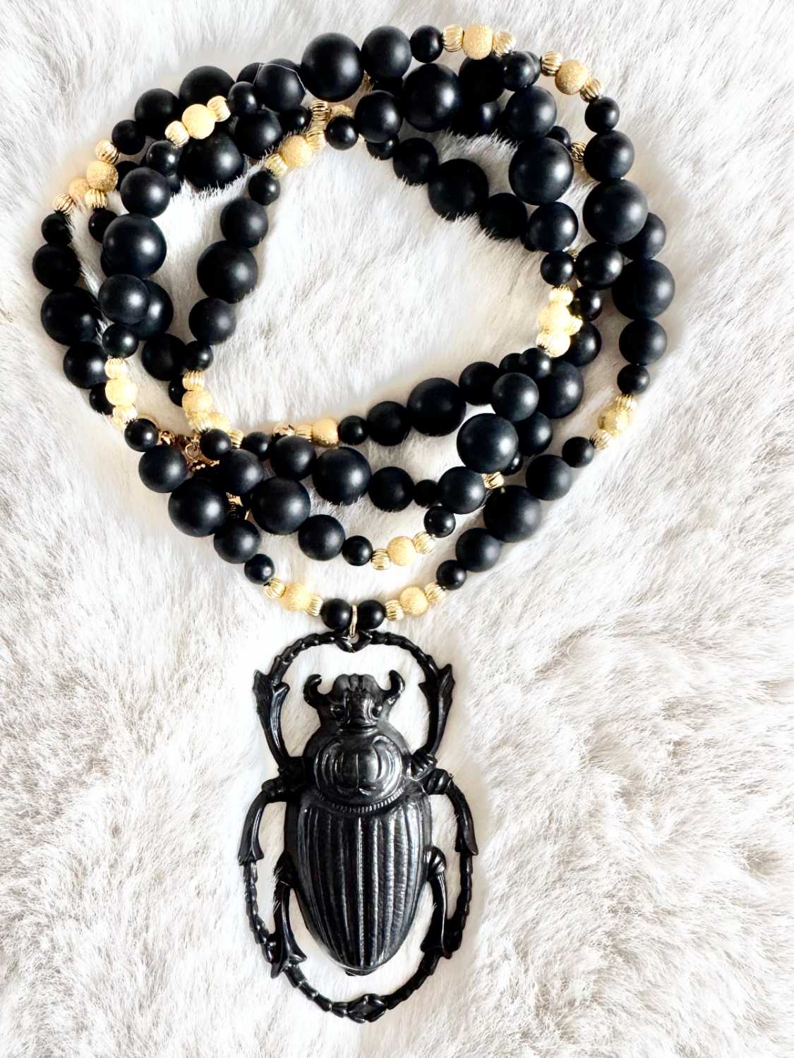 NOX-sautoir-scarabee-noir-metal-mat-perles-dorees-mademoiselle-oups Sautoir NOX en perles d’onyx noir mat de différentes tailles, perles dorées et grand pendentif scarabée noir, présenté sur fond clair.